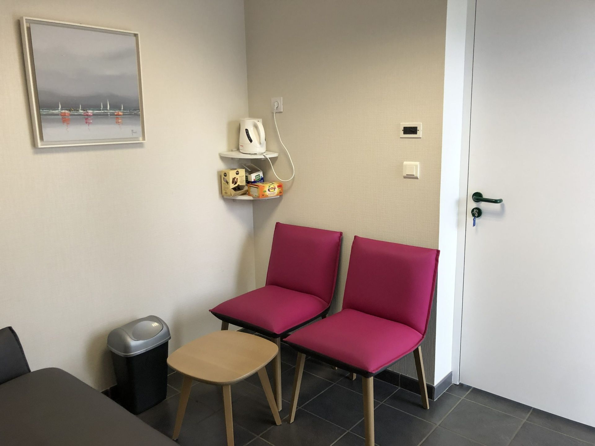 Petite salle d'attente avec deux chaises roses, une table d'appoint, une poubelle et une porte blanche.