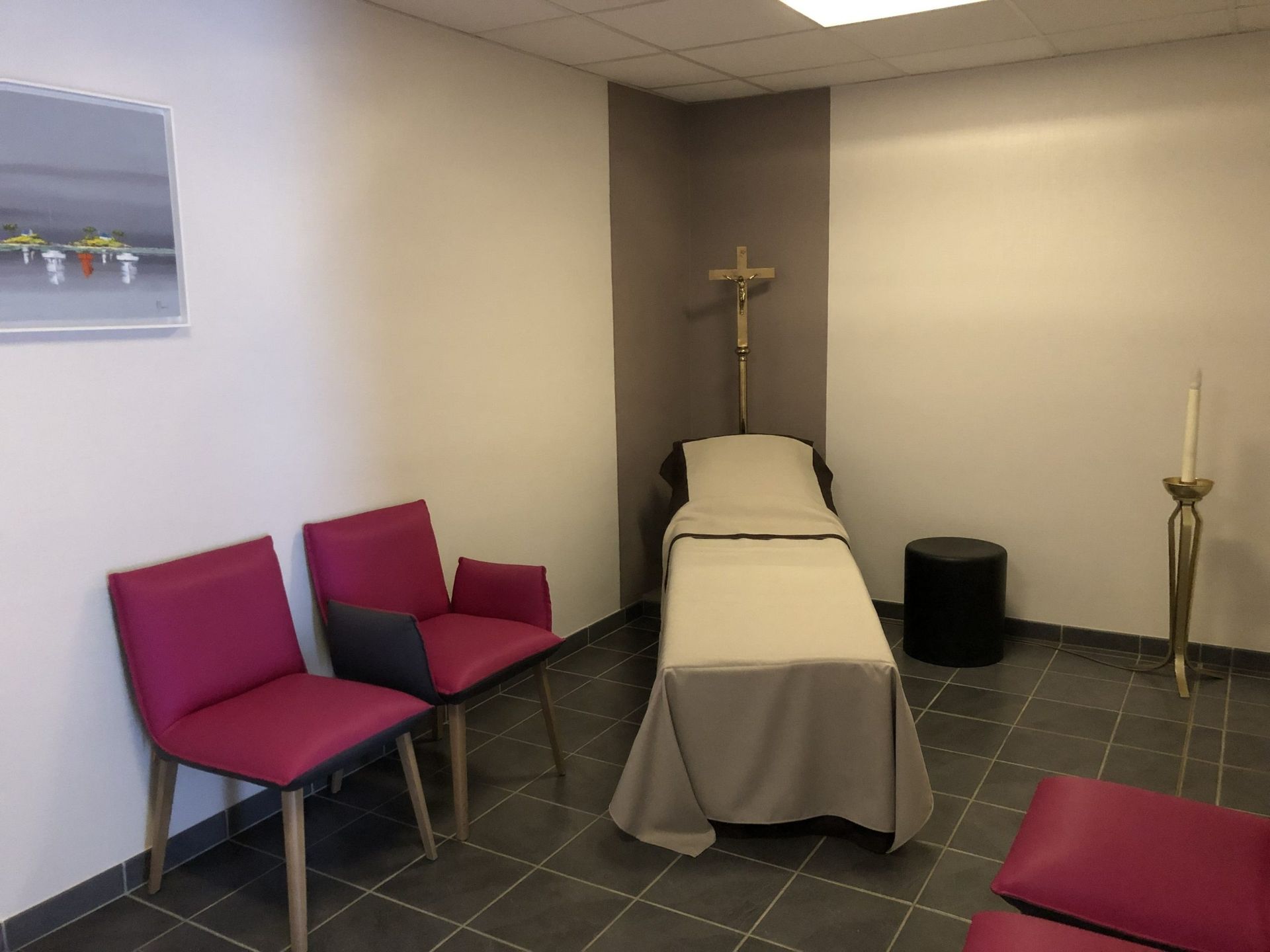 Chambre funéraire avec un lit, une croix et des chaises magenta sur un sol carrelé.