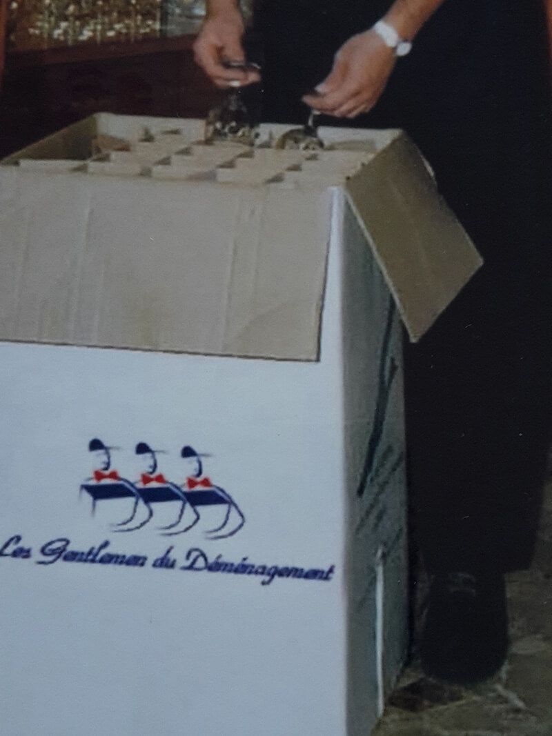 Personne déballant de la verrerie d'une boîte étiquetée « Les Gentilhommes du Déménagement » avec un logo représentant trois cygnes stylisés.