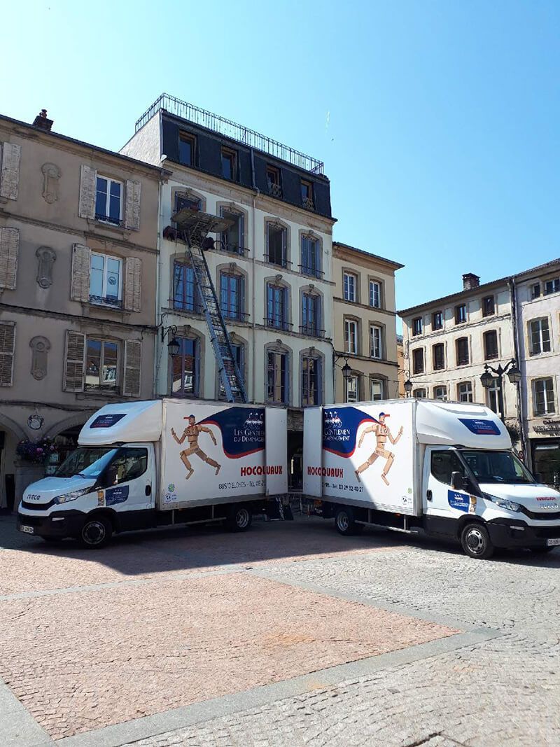 Deux camions blancs arborant des logos sont stationnés sur une place d'une ville européenne, avec des bâtiments en arrière-plan.