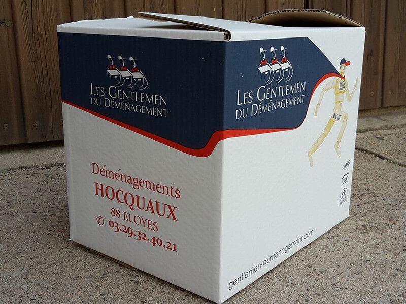 Boîte de déménagement avec le logo « Les Gentlemen du Déménagement » en blanc, bleu et rouge, sur du béton.