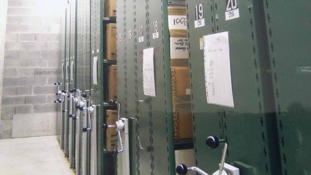 Des étagères métalliques vertes dans un débarras, remplies de boîtes d'archives, étiquetées avec des numéros et du papier.