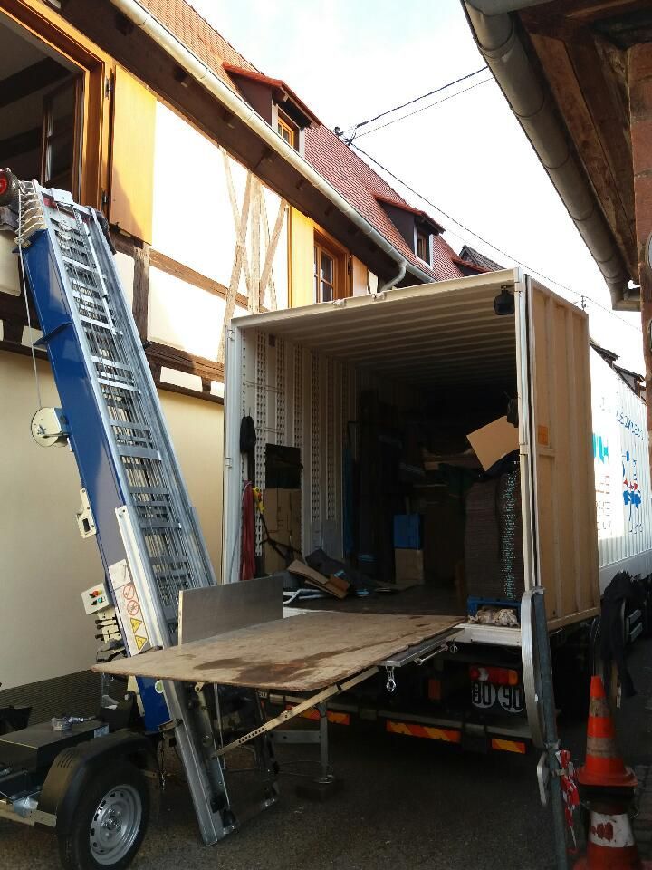 Un camion de déménagement avec hayon élévateur est appuyé contre un bâtiment ; on aperçoit du matériel et des fournitures de déménagement.