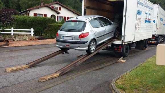Une voiture argentée est chargée sur un camion à l'aide de rampes en bois devant une maison.