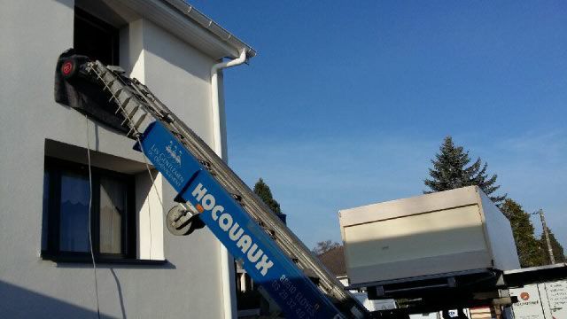 Un objet volumineux est hissé jusqu'à une fenêtre par un monte-charge bleu Hocquaux par une journée ensoleillée.