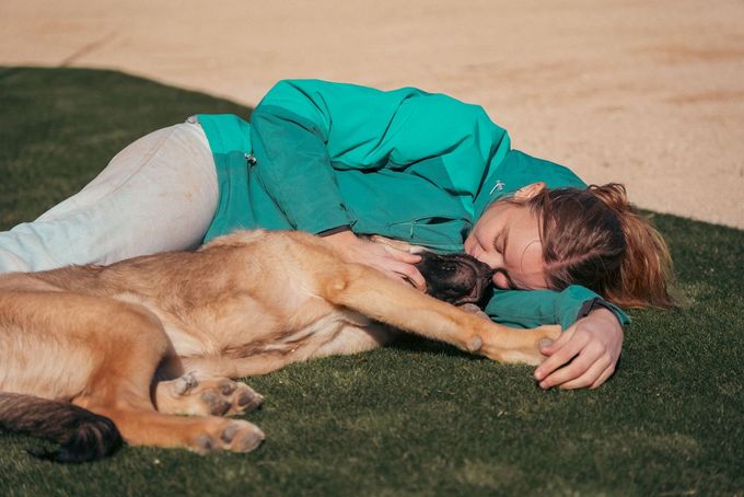 Una persona con una camisa color turquesa está tumbada en la hierba, abrazando a un perro de color canela al aire libre.