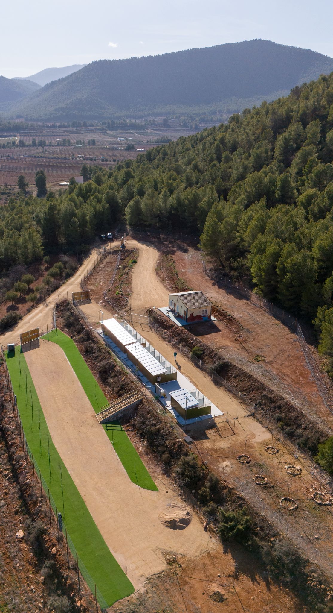 Vista aérea de una obra en construcción de un camino de tierra junto a una ladera verde, con montañas al fondo.