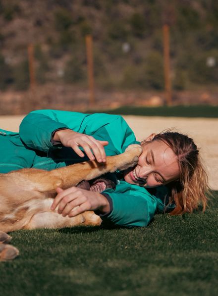 Una persona con una chaqueta turquesa abraza a un perro dorado sobre el césped al aire libre.