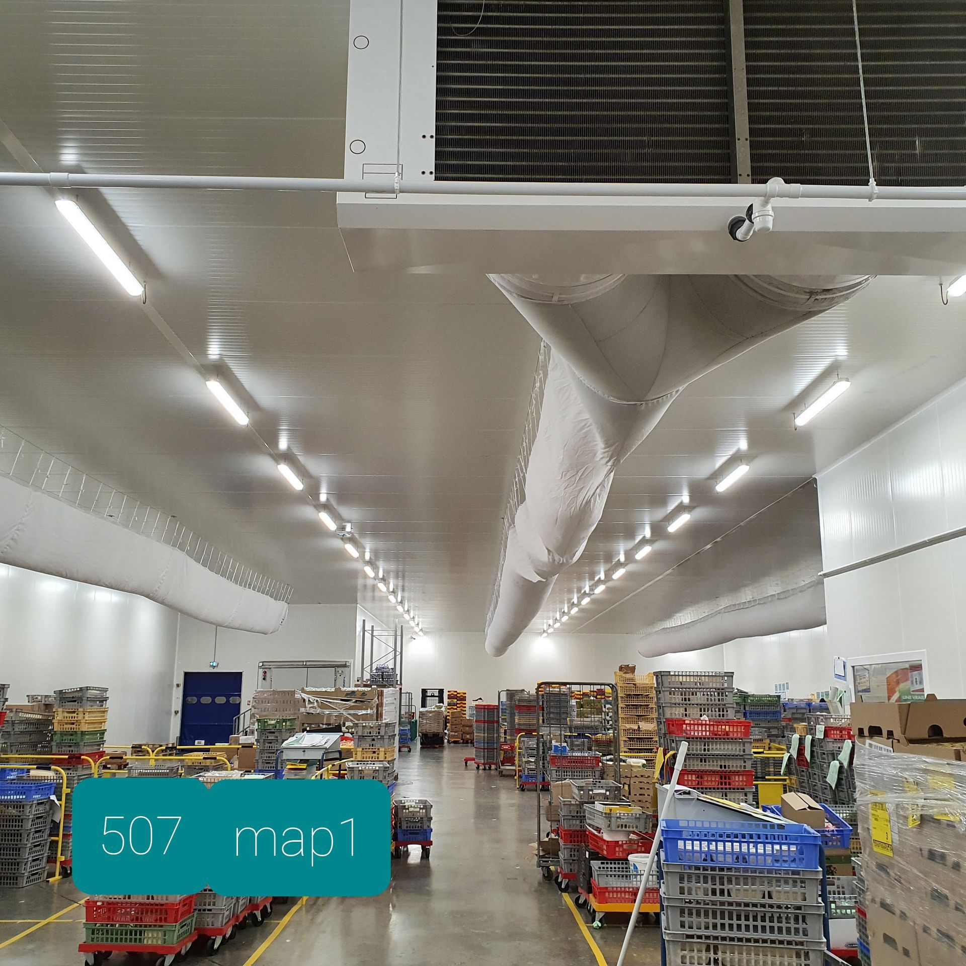 Allée lumineuse d'entrepôt avec des étagères, des marchandises empilées et de grandes grilles d'aération blanches au plafond.