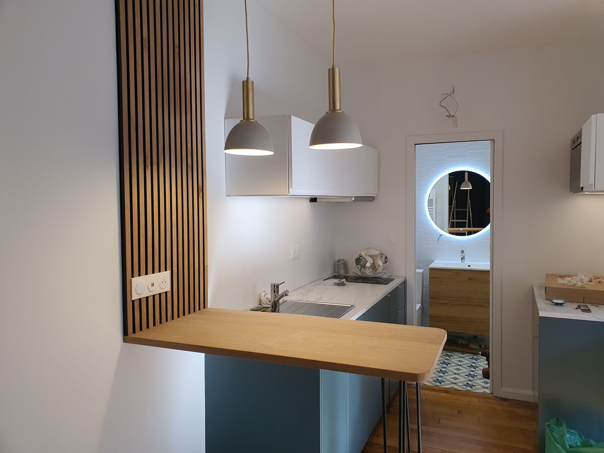Cuisine moderne avec bar en bois, suspensions lumineuses et miroir reflétant la pièce adjacente.