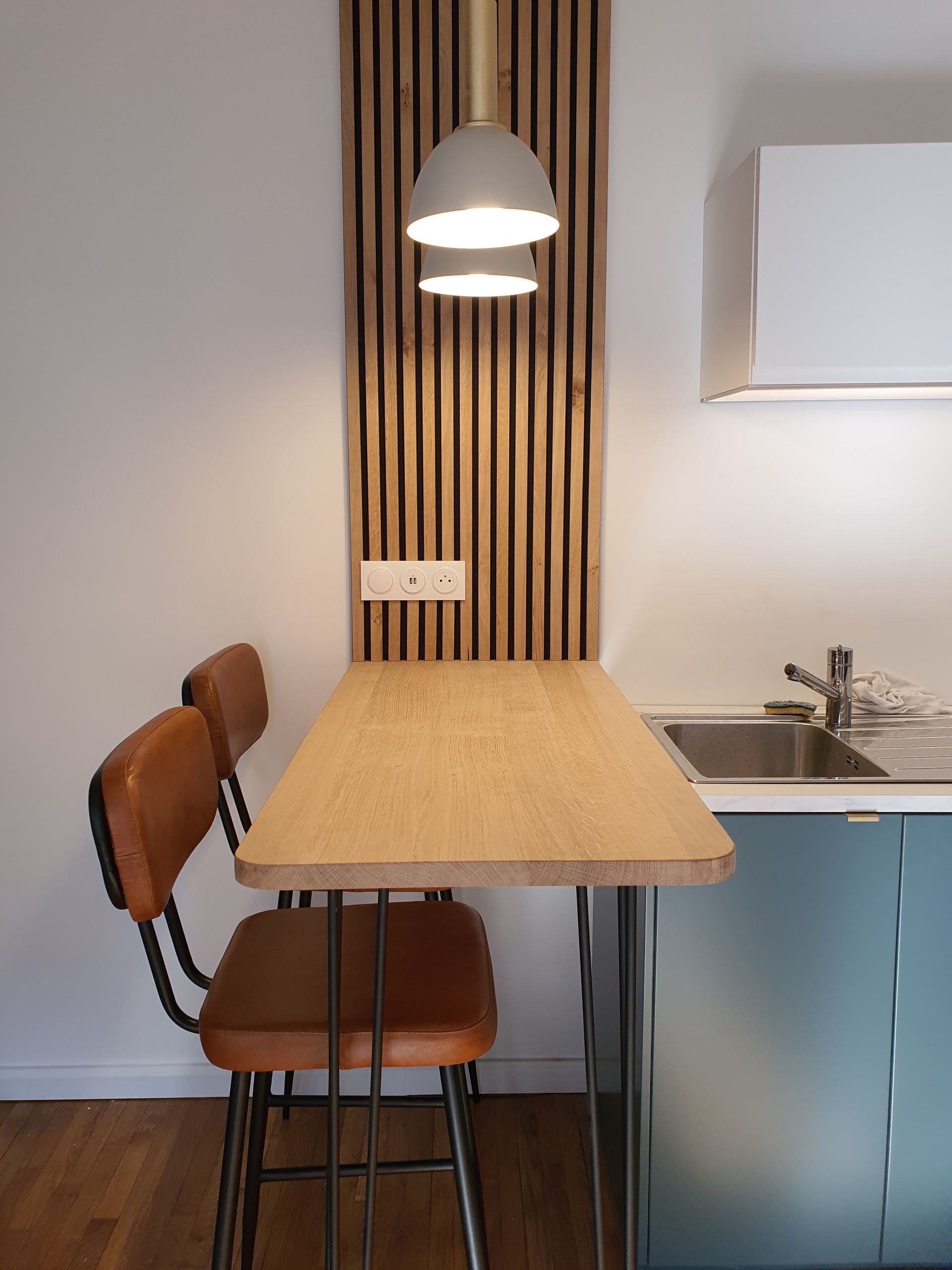 Petit coin cuisine avec table en bois, deux chaises, suspension lumineuse et lambris mural.
