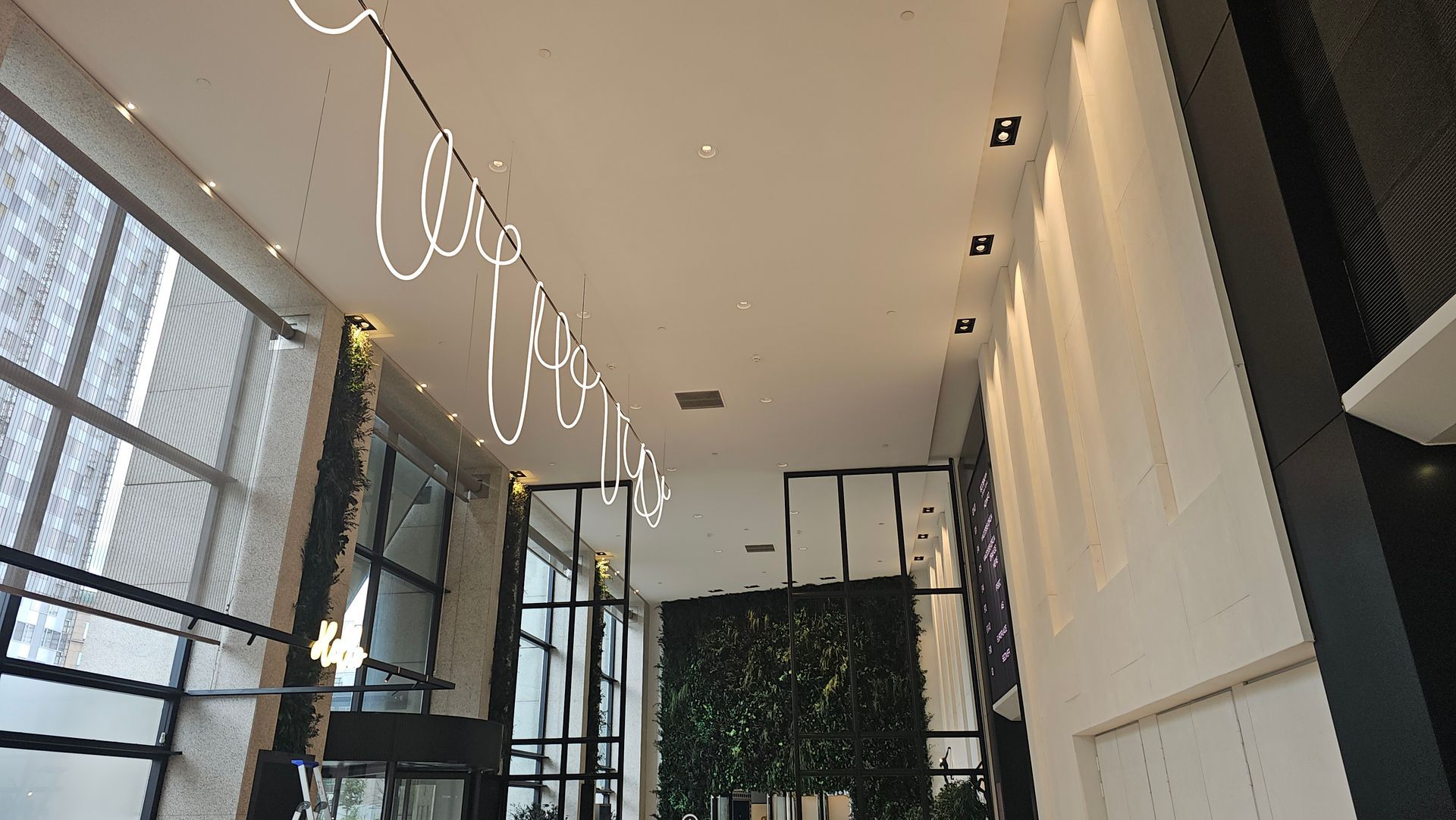 Hall d'entrée moderne avec de hautes fenêtres, des luminaires suspendus et un mur végétal sous un haut plafond.
