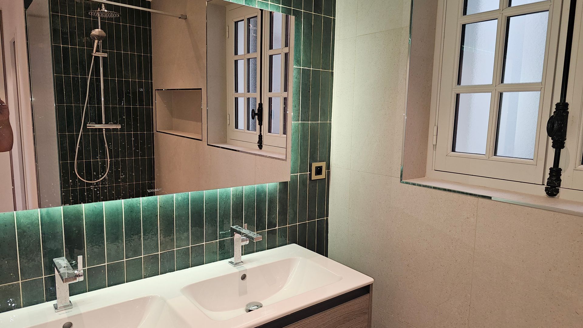 Meuble en bois avec double vasque, crédence en carrelage vert, miroir rectangulaire éclairé.