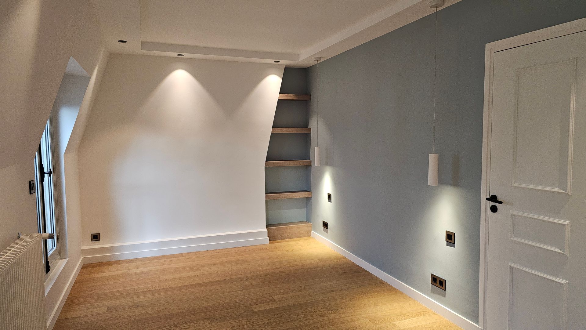 Pièce vide avec parquet clair, murs gris, porte blanche et étagères murales en bois.