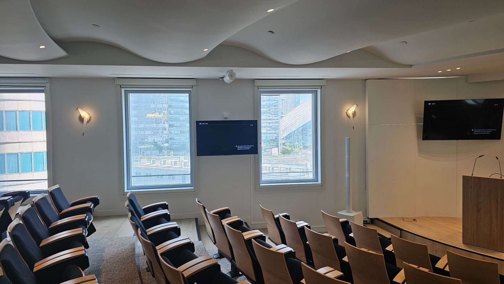 Salle de conférences avec rangées de chaises, écrans muraux et fenêtres donnant sur la ville.
