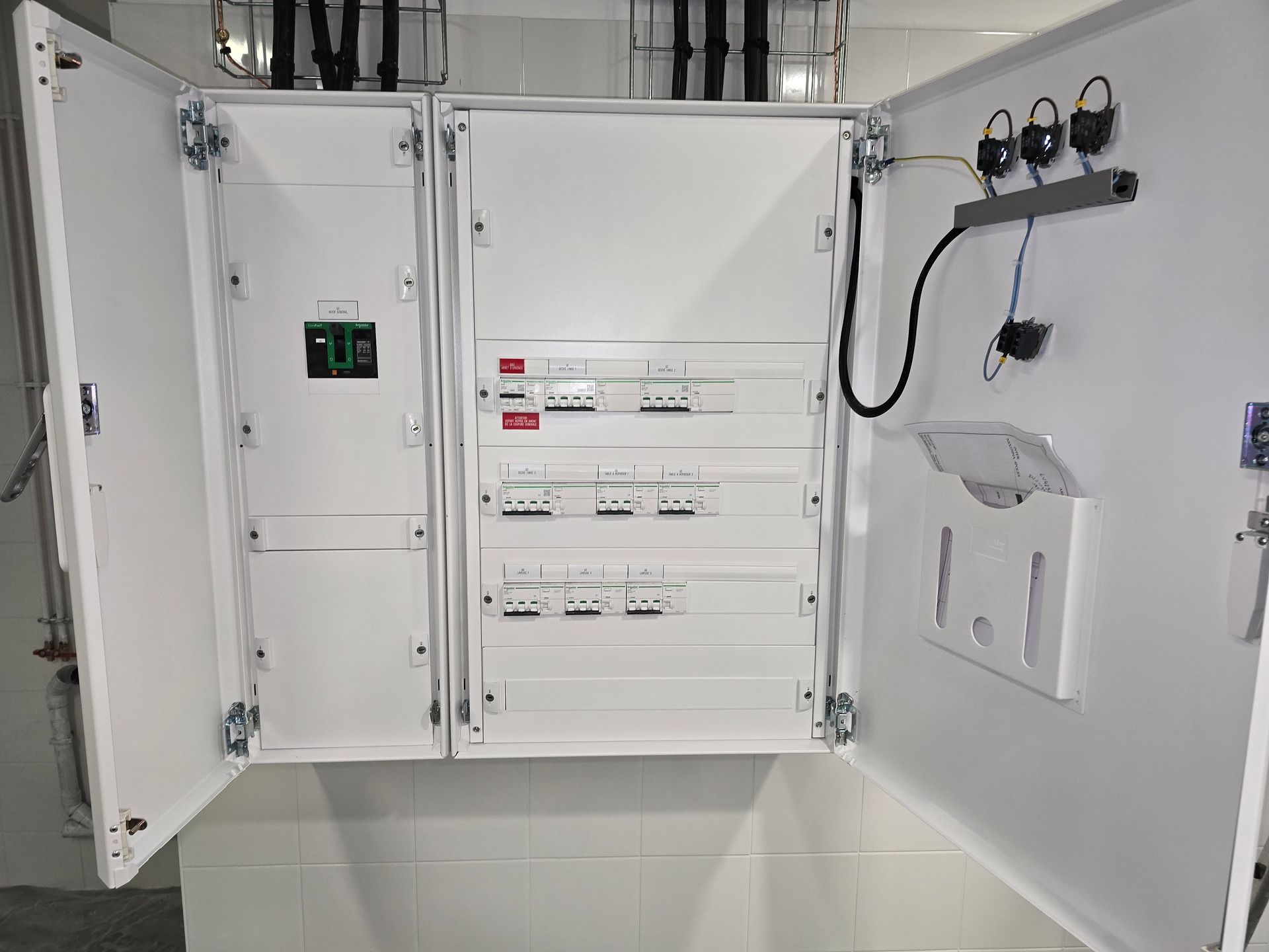 Panneau de commande électrique ouvert avec interrupteurs, compteurs et câblage à l'intérieur d'une armoire blanche.