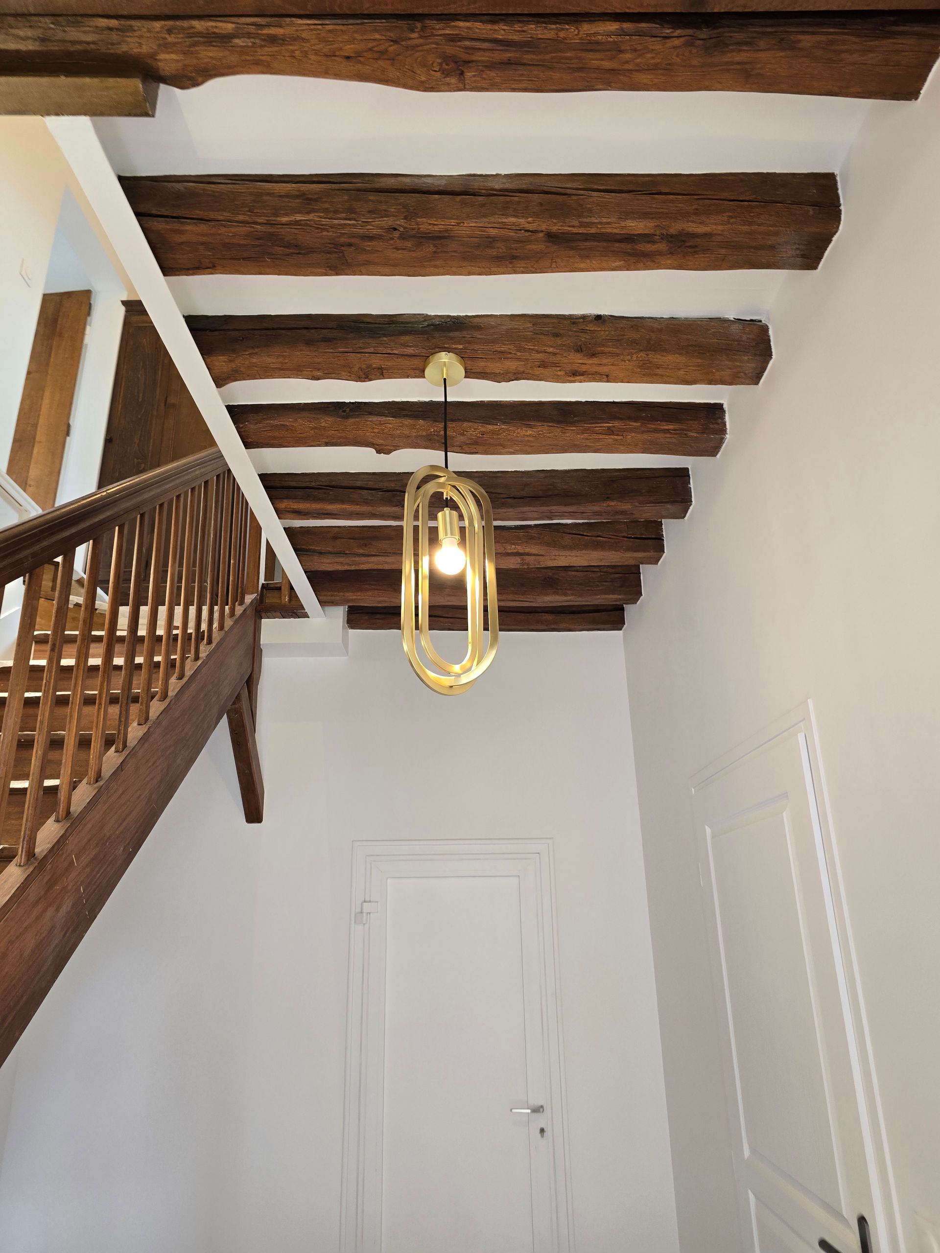 Escalier étroit avec poutres en bois, murs blancs et suspension lumineuse dorée au-dessus de deux portes blanches fermées.