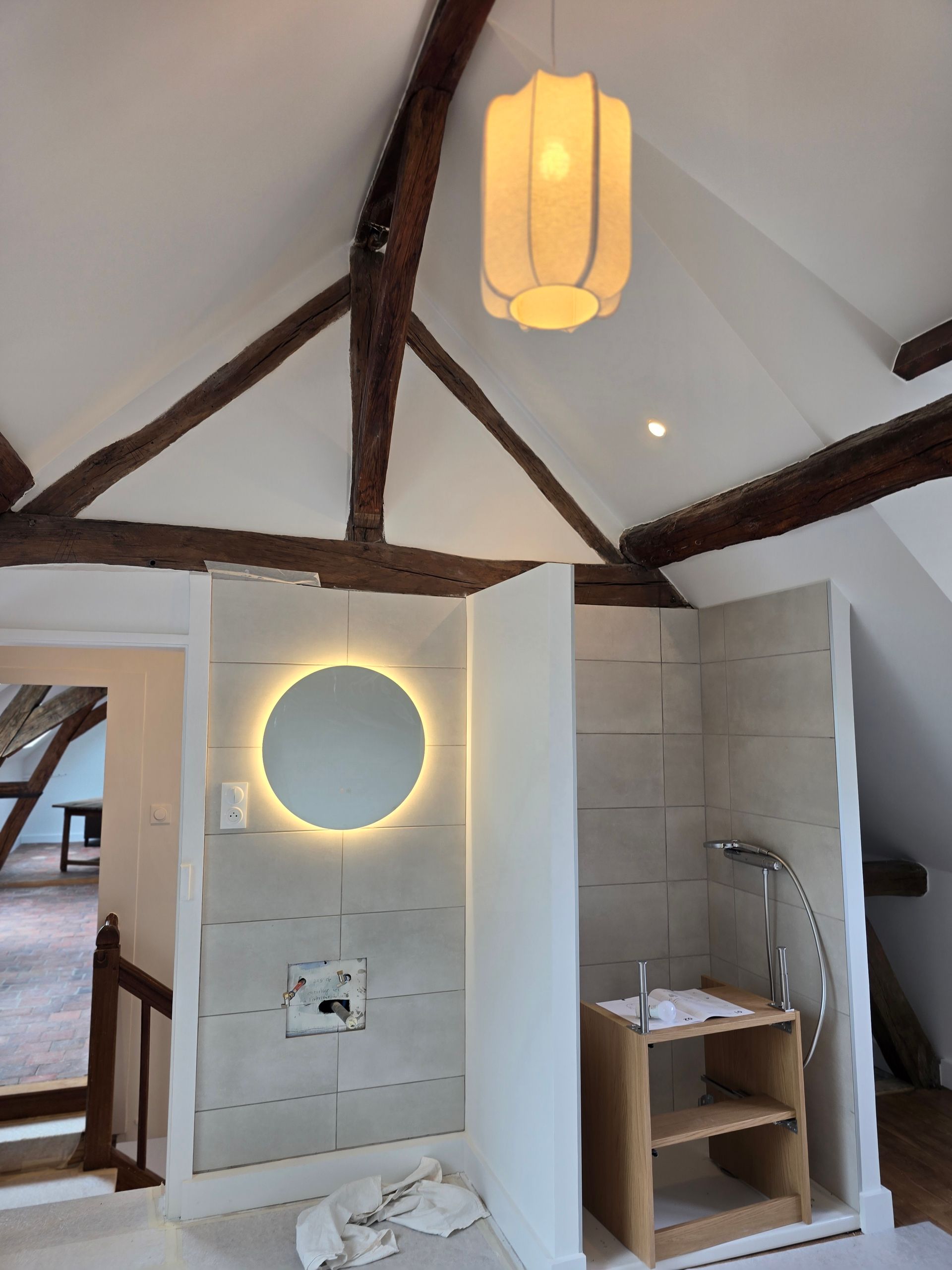 Salle de bains mansardée en travaux, avec poutres en bois, miroir rond éclairé.