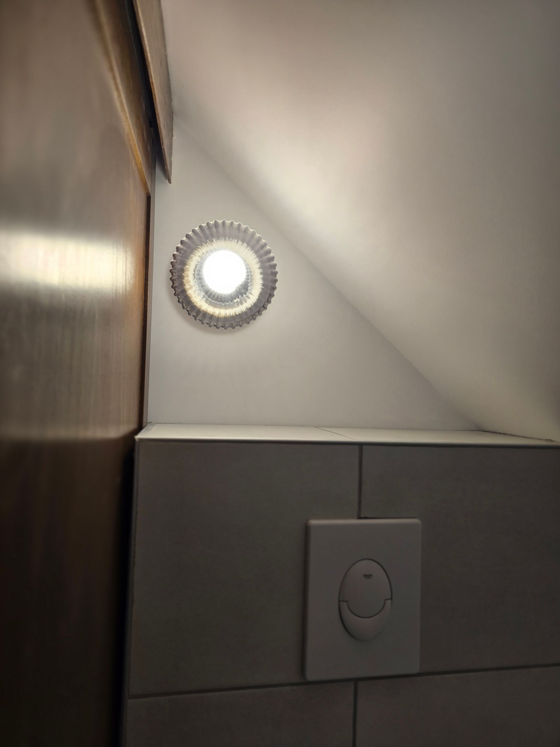 Petit luminaire mural circulaire avec éclairage diffus installé dans un angle au-dessus d’un interrupteur.