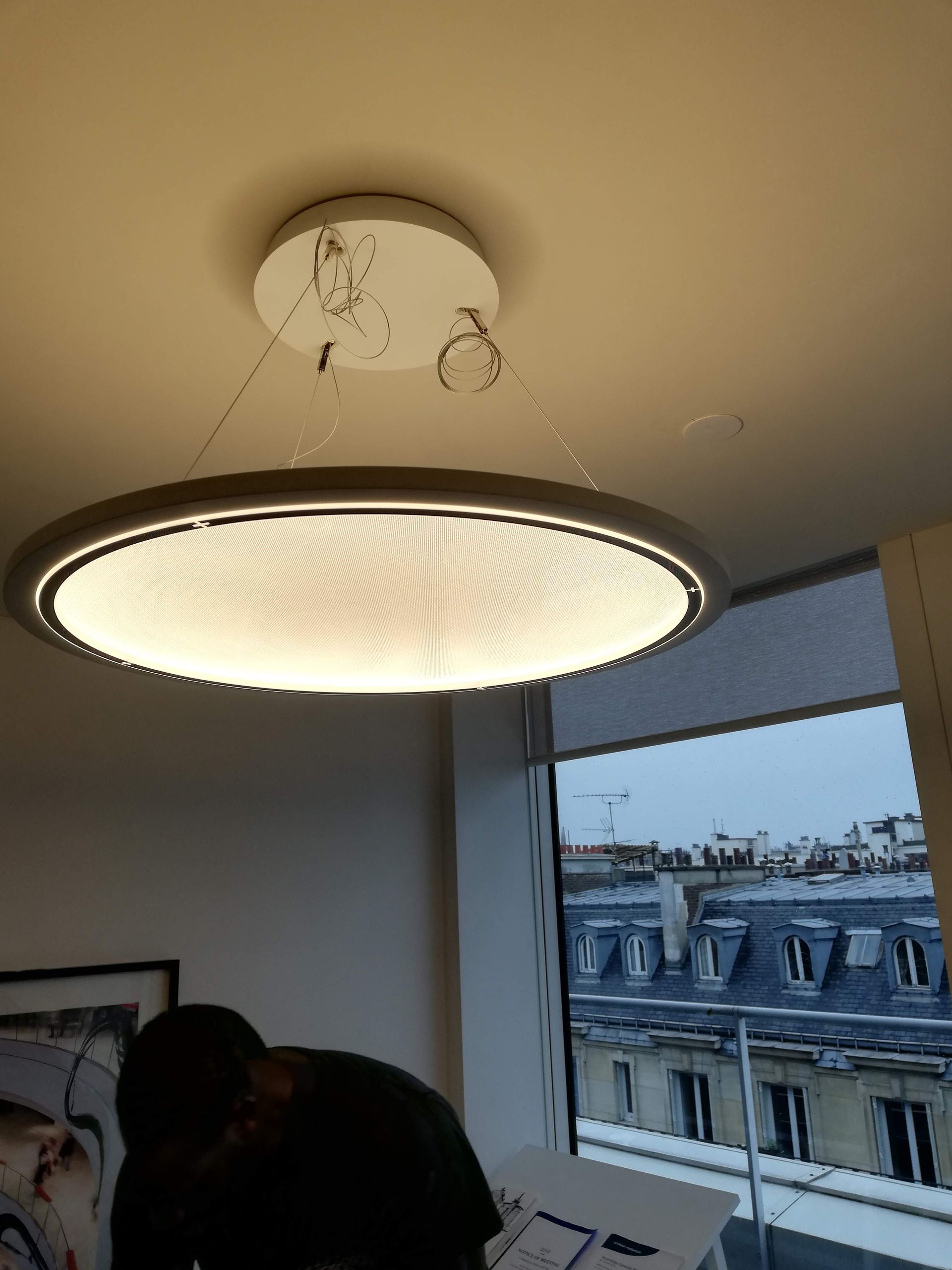 Lampe au design circulaire moderne, suspendue à un plafond blanc, près d'une fenêtre avec vue sur les toits de Paris.