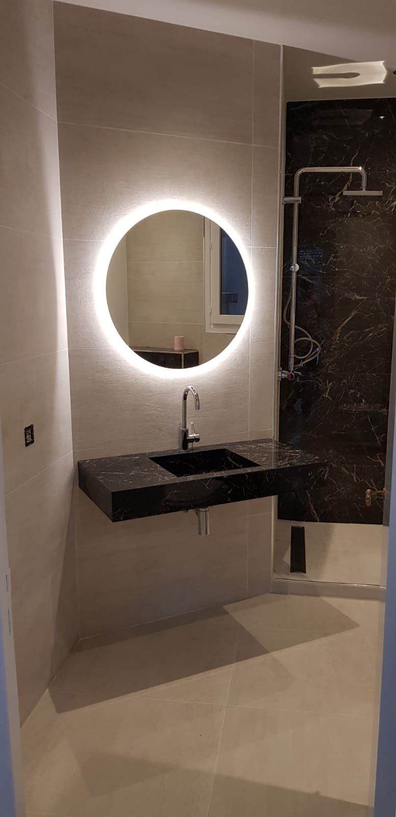 Salle de bains moderne avec miroir rond rétroéclairé, vasque noire et douche effet marbre.