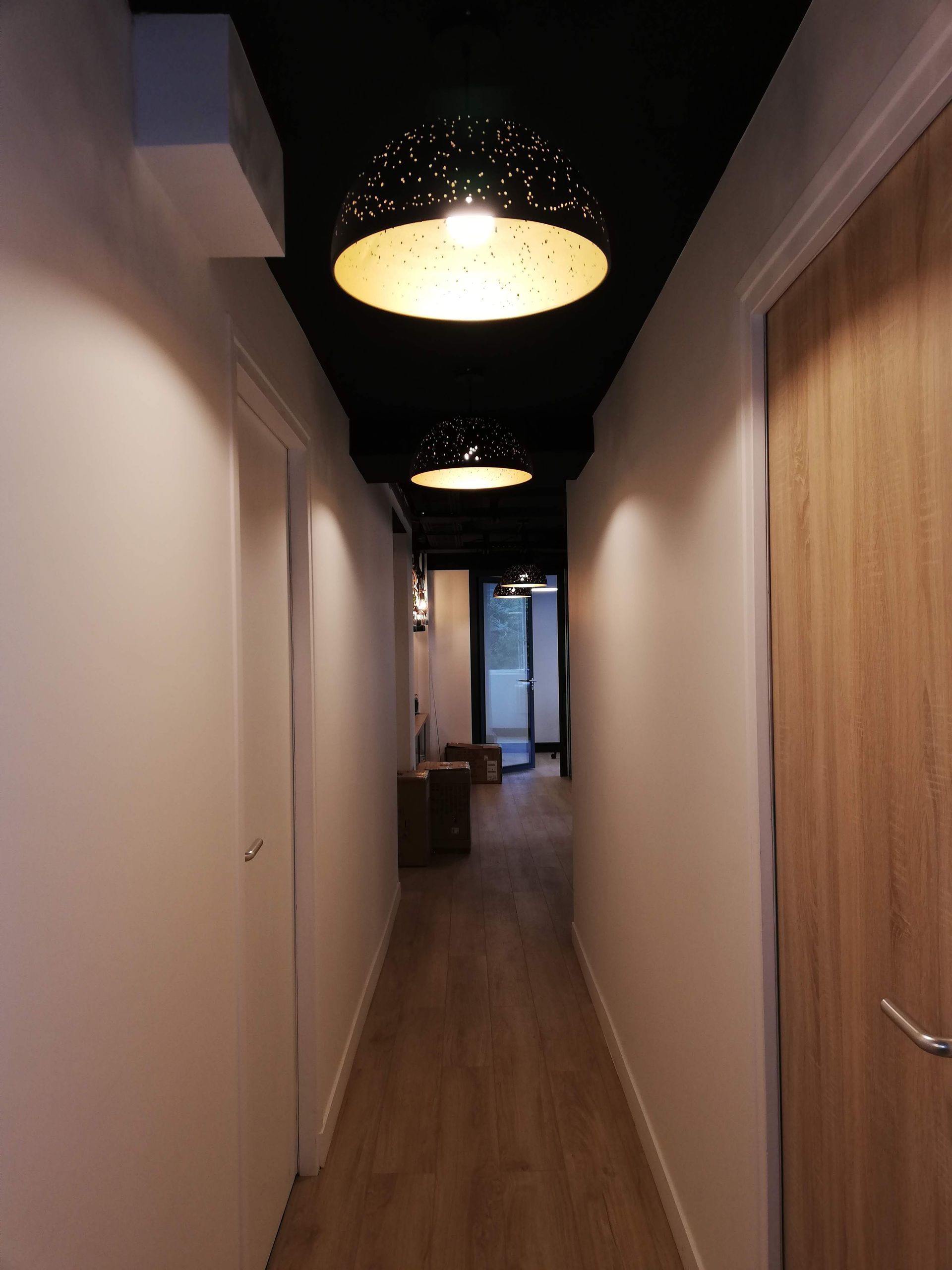 Long couloir avec parquet, murs blancs et plafonniers ronds noirs.