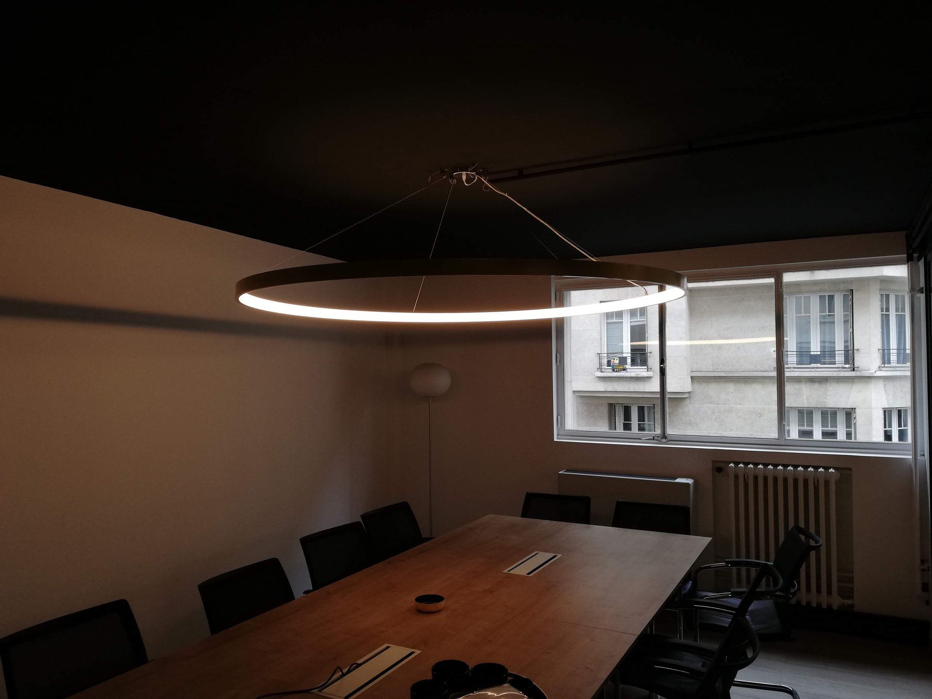 Salle de conférence sombre avec une longue table en bois, des chaises noires et un plafonnier circulaire.