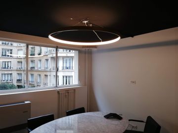 Salle à manger avec table ronde effet marbre, plafonnier moderne et grande fenêtre donnant sur des immeubles.