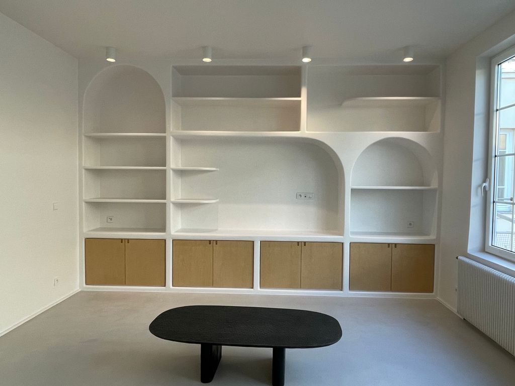 Étagère murale blanche intégrée avec placards en bois, table ovale noire et fenêtre dans une pièce lumineuse.