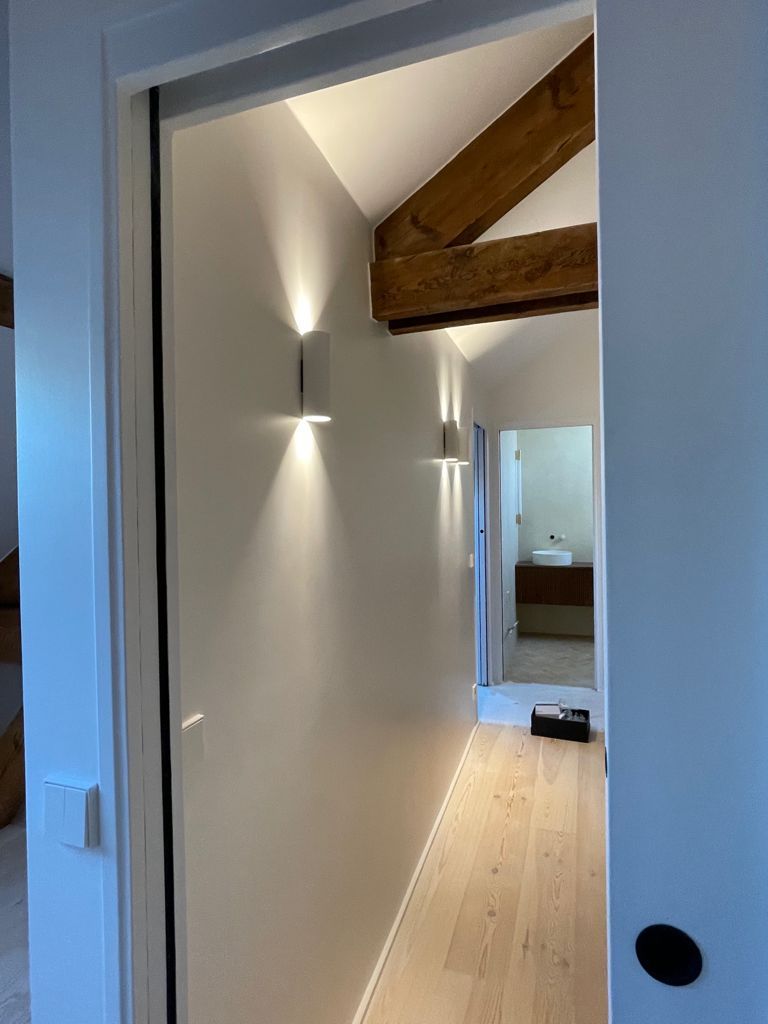 Couloir étroit avec parquet clair, murs blancs, poutre apparente et appliques murales allumées.