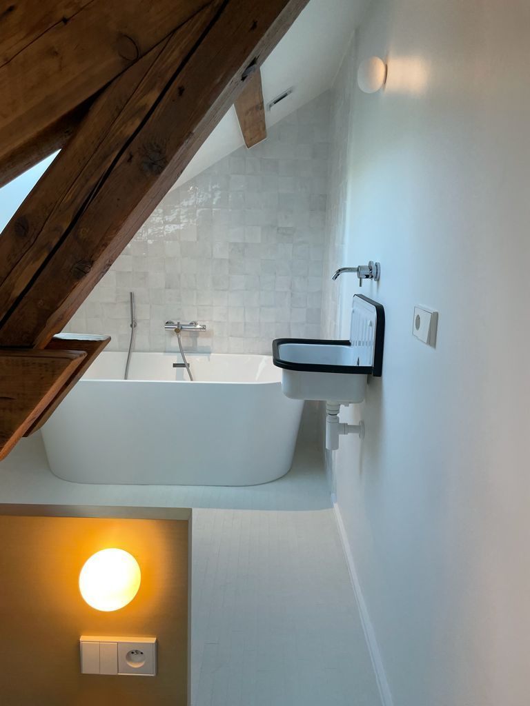 Salle de bains mansardée avec poutres apparentes, baignoire îlot, mur carrelé blanc.