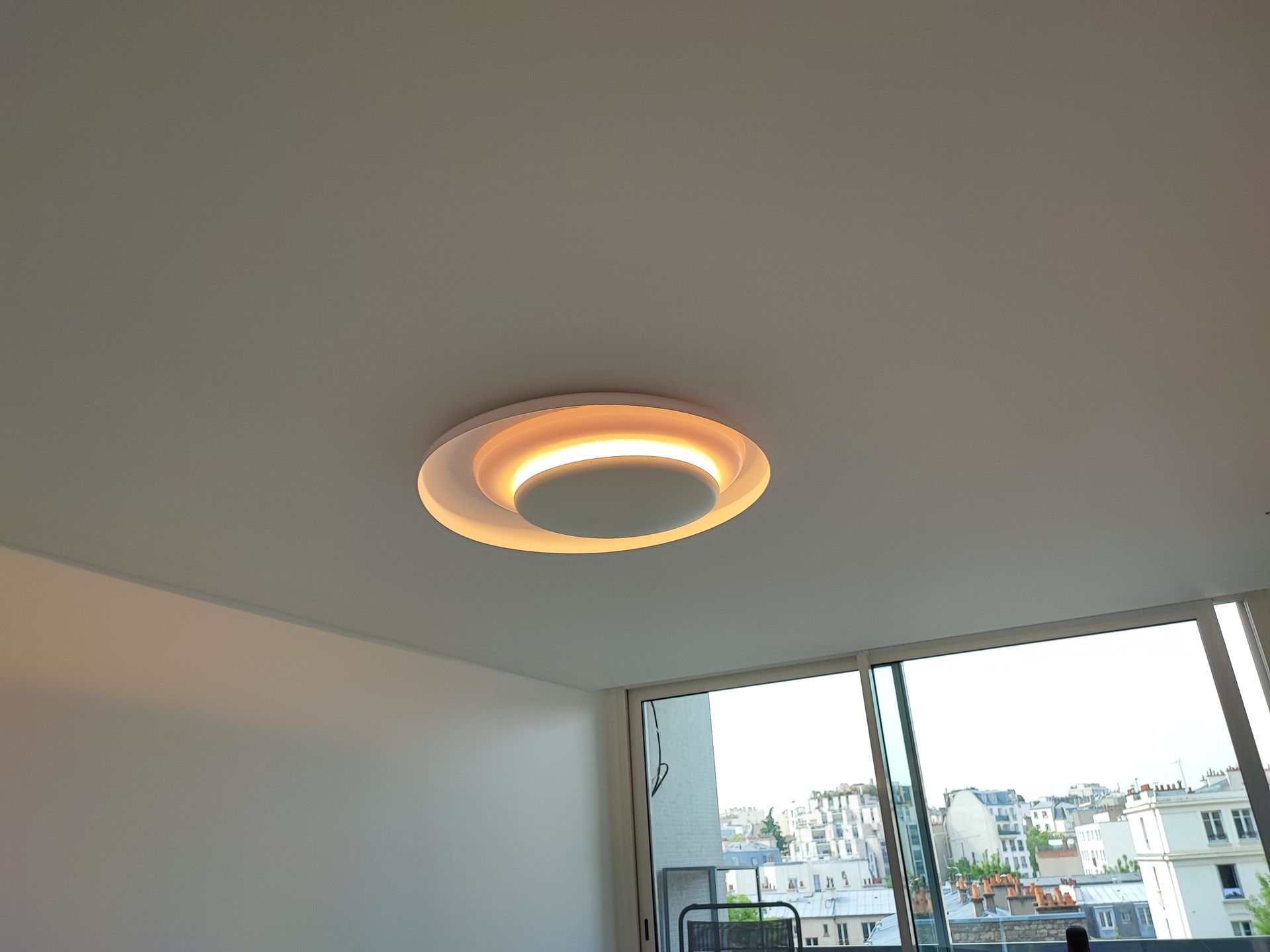 Plafonnier moderne dans une pièce avec une grande fenêtre et vue sur les toits de Paris.