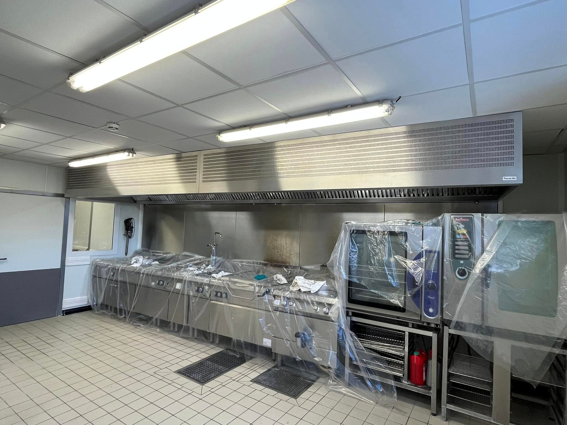 Une cuisine professionnelle équipée de postes de préparation recouverts de film plastique sous une hotte aspirante.