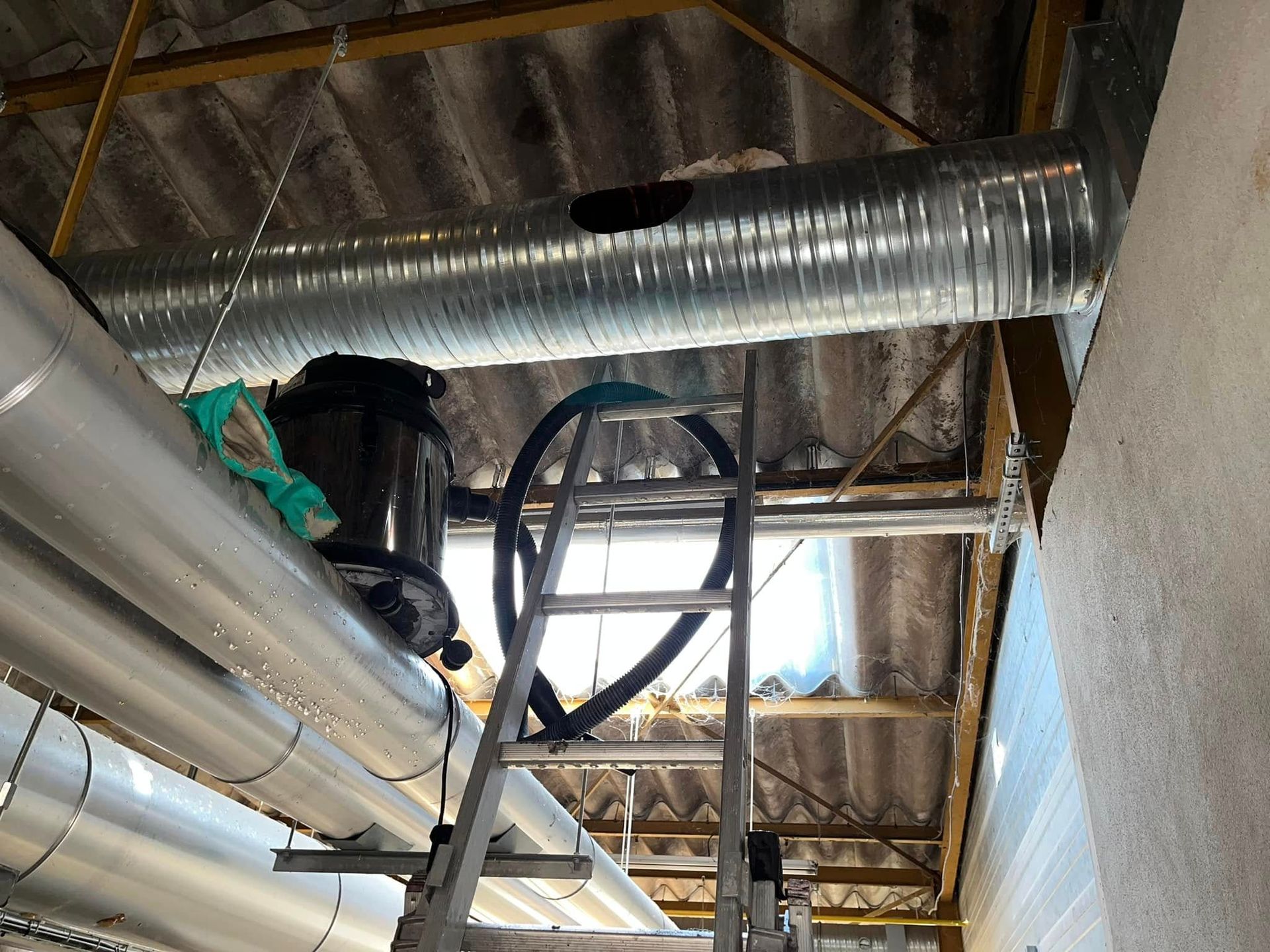 Une échelle métallique se dresse sous de grands conduits de ventilation industriels suspendus à un plafond ondulé.