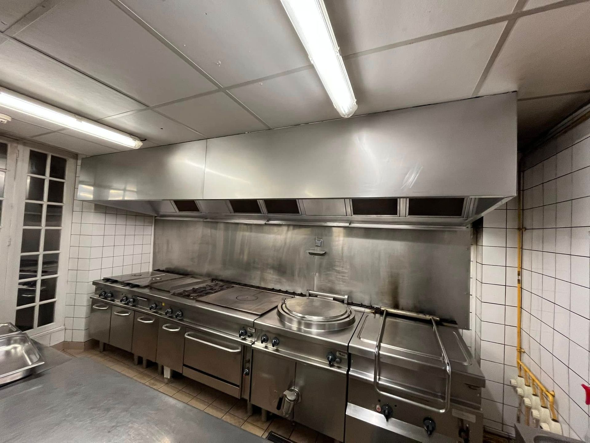 Une cuisinière industrielle en acier inoxydable, surmontée d'une grande hotte aspirante.