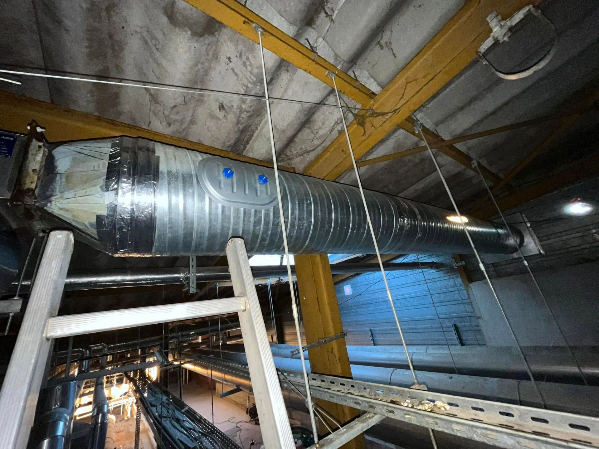 Un long conduit cylindrique argenté est suspendu à des poutres en acier jaune au-dessus d'une passerelle métallique.