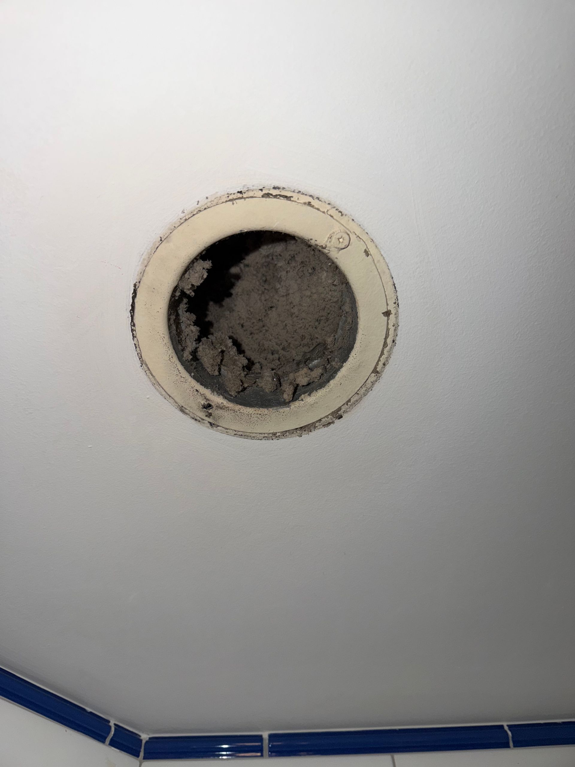 Une grille d'aération ronde et blanche au plafond, recouverte d'une épaisse couche de poussière grise et de peluches.