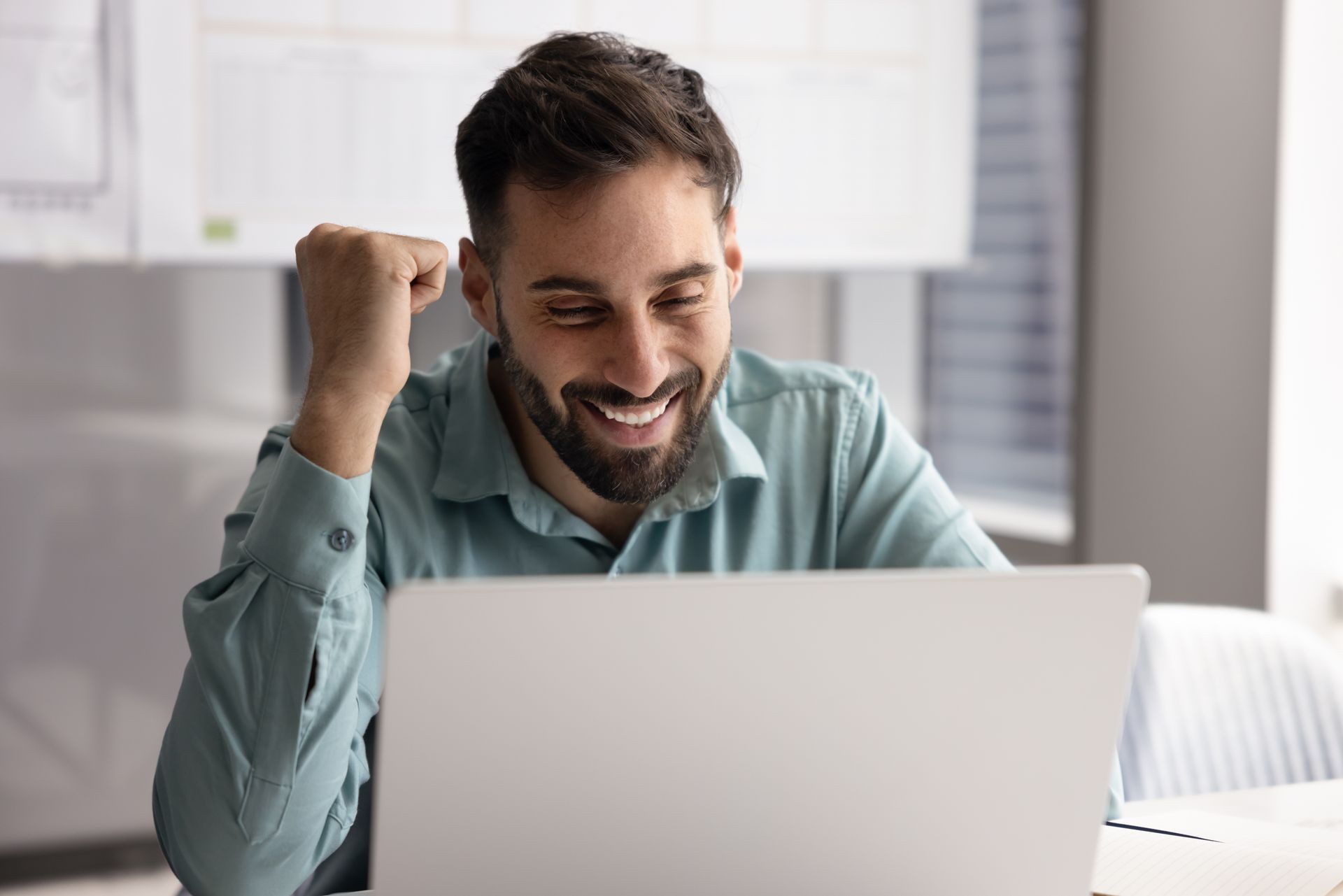 Un homme souriant, les yeux rivés sur son ordinateur portable, célèbre sa victoire, le poing levé. À l'intérieur, dans un bureau.