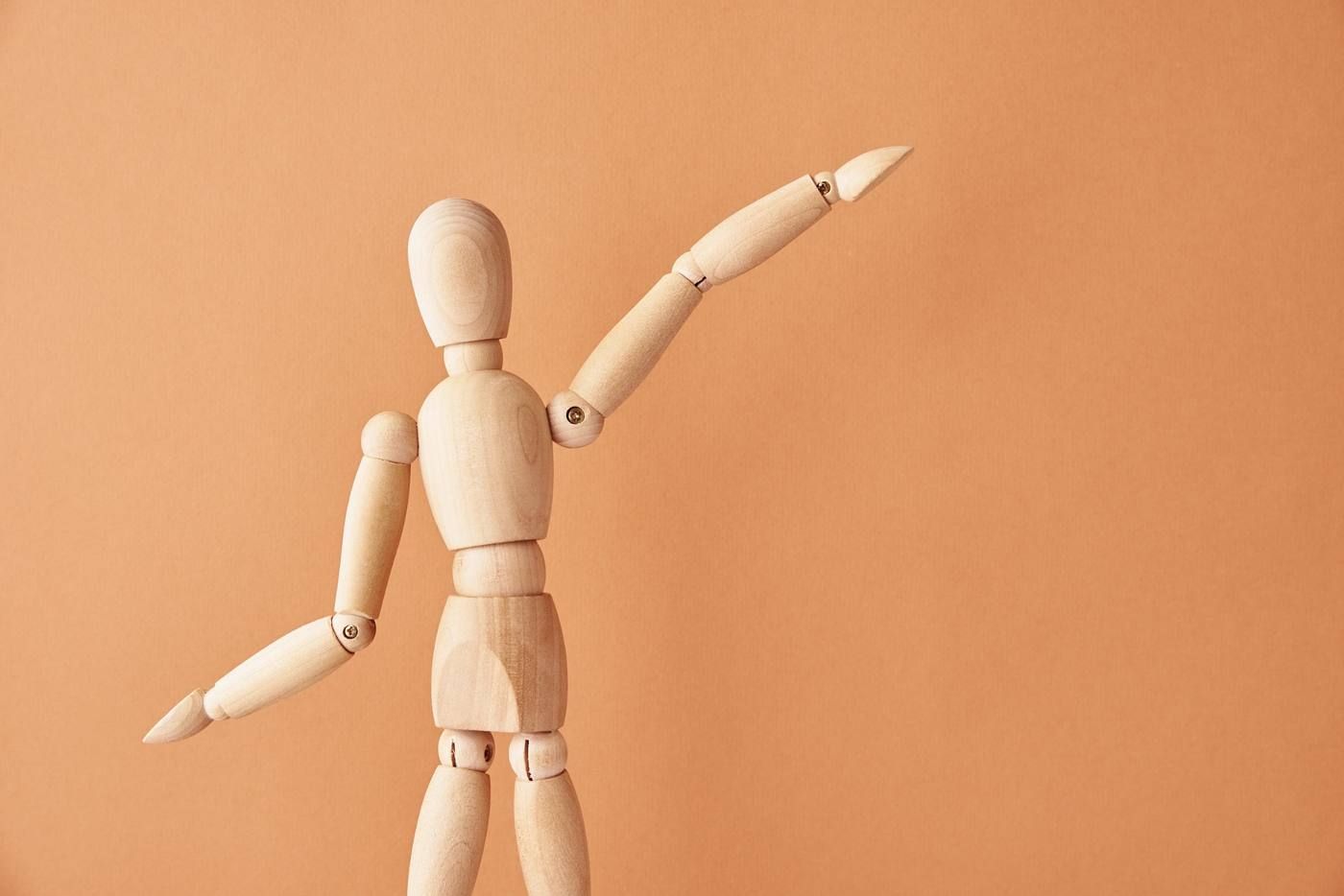 Maniquí de madera de artista posando con un brazo levantado sobre un fondo color melocotón.