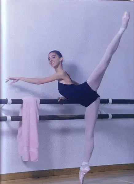 Bailarina de ballet, con leotardo oscuro, practicando una extensión de pierna alta en la barra de un estudio de danza.