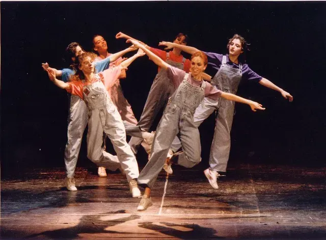 Bailarines con overoles azul claro realizan una coreografía sobre un escenario con fondo oscuro.