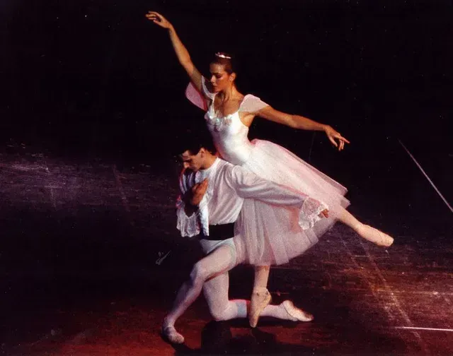 Bailarines de ballet actuando, una mujer elevada, un hombre arrodillado; escenario con fondo oscuro, foco.
