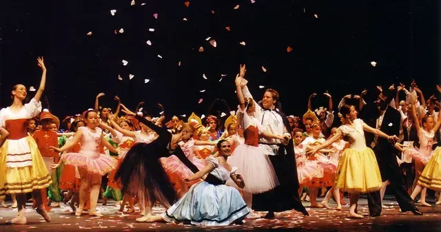 Bailarinas de ballet con coloridos trajes en el escenario, confeti cayendo, ambiente festivo.