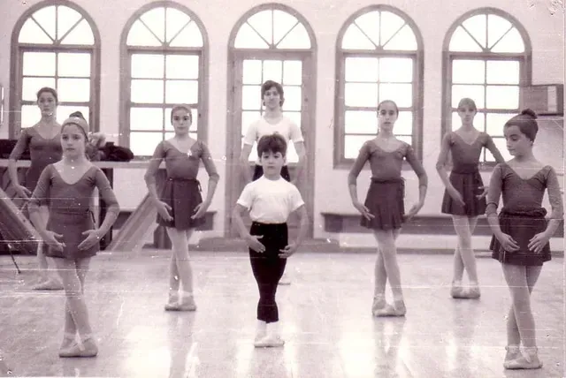 Clase de ballet con instructor; estudiantes con leotardos y tutús en un estudio con grandes ventanas.
