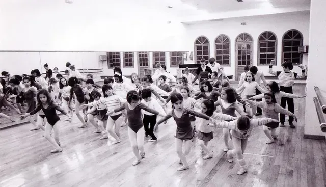 Niños en una clase de baile, practicando posturas en un estudio luminoso con barra y pared espejada.