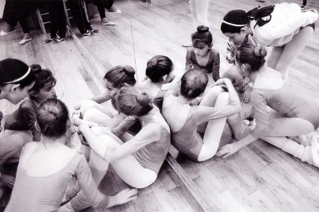 Jóvenes bailarinas con leotardos sentadas en círculo sobre un suelo de madera, algunas sujetando sus piernas, otras personas al fondo.