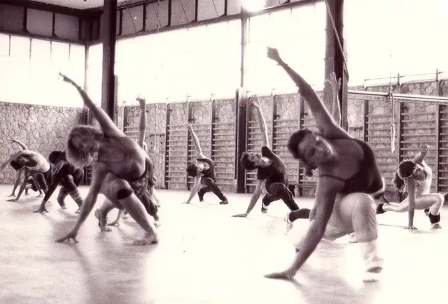 Bailarines en un estudio, realizando una secuencia similar al yoga con los brazos en alto y los cuerpos inclinados. Paredes con barras de escalada. Blanco y negro.