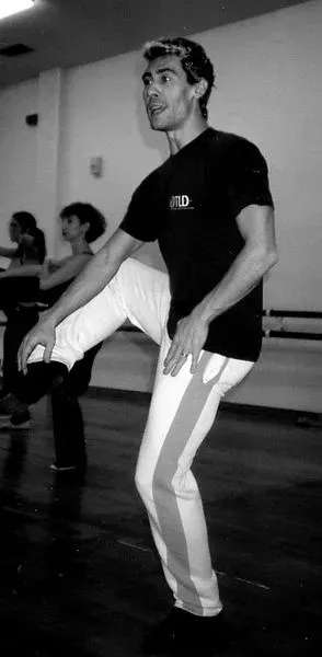 Hombre en un estudio de danza, levantando la pierna. Lleva una camisa negra, pantalones blancos a rayas y está concentrado en el movimiento.