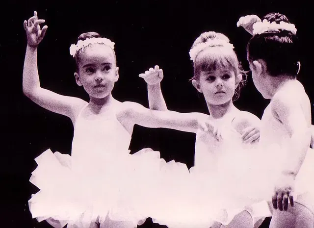 Tres jóvenes bailarinas con tutús blancos y diademas actuando en el escenario con los brazos en alto.