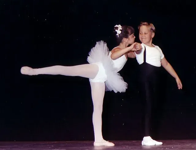 Una niña con tutú se mantiene en equilibrio, un niño con pantalones negros y tirantes le sujeta la mano, en el escenario.
